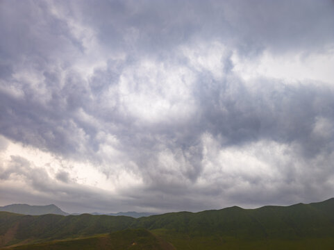 Landscape photograph captures rolling green hills beneath a dramatic, cloudy sky. Heavy grey clouds contrast with lighter patches, creating an atmospheric scene.