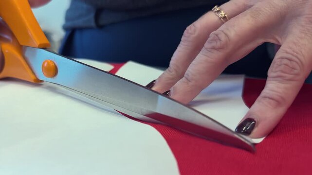 A woman with manicured nails uses scissors to cut paper, shaping a design on a red fabric surface. The workspace is organized with various crafting materials visible in the background