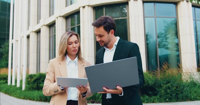 Close-up of a businessman with a laptop and an elegant young businesswoman using a smartphone outside a modern office building, discussing ideas and analyzing data together.