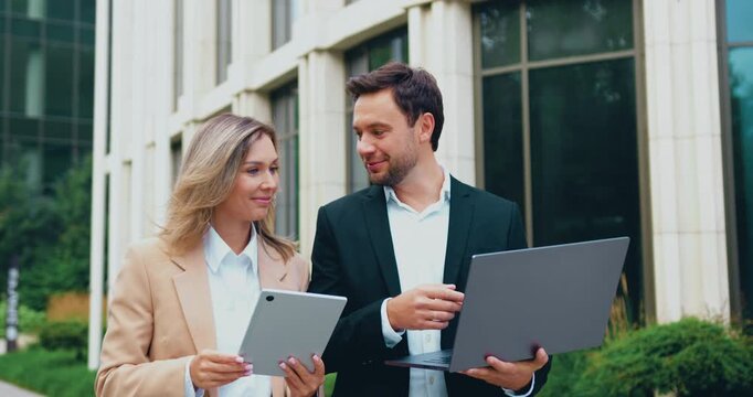 Confident businessman with laptop and elegant businesswoman with tablet walking together down the street toward a modern office building, discussing work and collaborating on a project