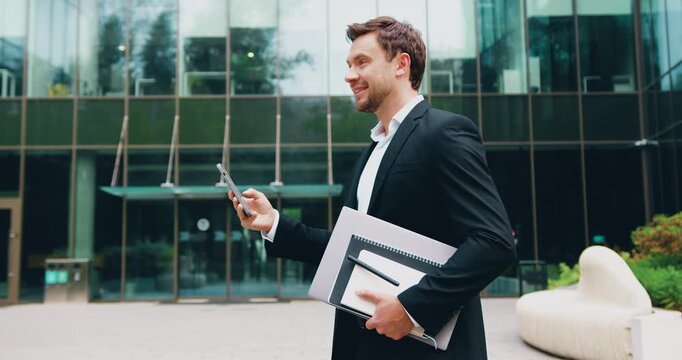 Confident businessman in a formal suit using smartphone for online communication while walking outdoors near modern office building
