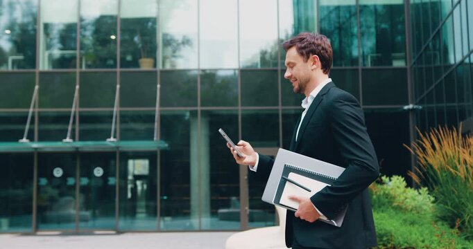 Confident Caucasian man in jacket and tie walking outdoors on a sunny summer day, using a smartphone and typing a message with a modern glass building behind him