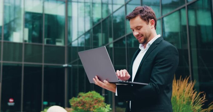 Handsome businessman working on a laptop outdoors near a modern glass office building, representing remote work, digital communication, and a modern urban business lifestyle