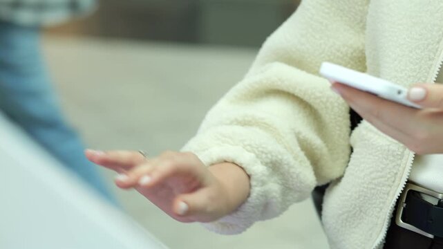 Person holds phone, gestures with free hand. Wears cream-colored fleece jacket and dark pants. Background blurred, indoor setting. Hands in focus, neutral expression
