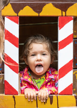 Playful generation alpha child sticks out her tongue through a colorful wooden playhouse window. Funny candid portrait captures childhood humor, curiosity, spontaneity, and carefree play.