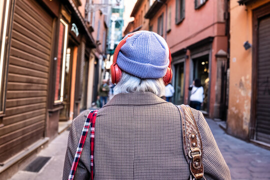Generation x traveler wearing red headphones and knit beanie walks through a narrow old town street. Rear view highlights independent urban travel, music, fashion, and everyday city lifestyle.