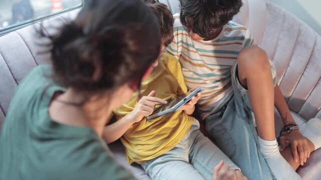 Mother teaching her two young sons how to use a tablet computer while sitting together on the couch, showing them educational apps and games for their development