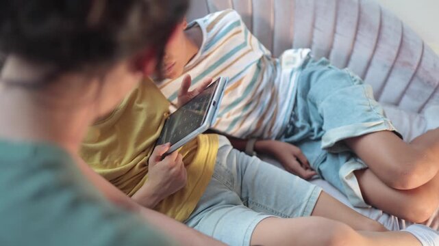 Over the shoulder view of young brother sleeping while his sibling is using a tablet, relaxing together on a cozy couch at home during a sunny afternoon