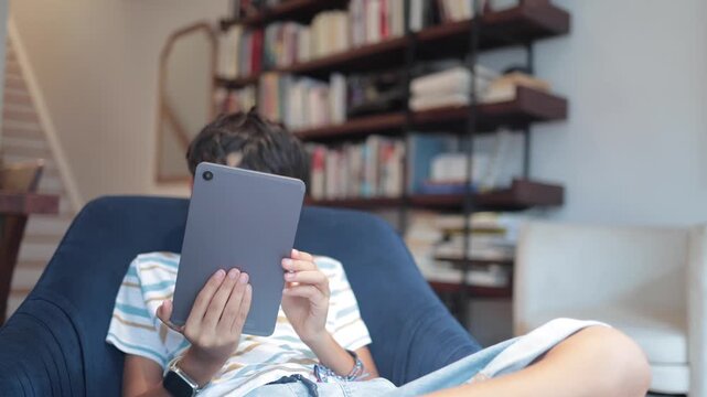 Young student learning and playing games on a tablet, sitting comfortably in an armchair at home with a large library in the background, promoting digital education