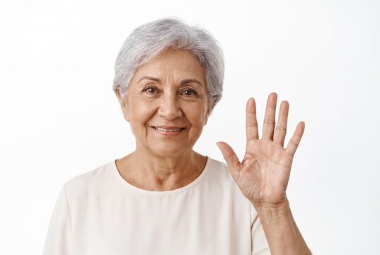  Retrato de una amigable mujer mayor espa&ntilde;ola sonriendo y saludando con la mano. Feliz abuela hispana con canas mirando a la c&aacute;mara sobre fondo blanco aislado