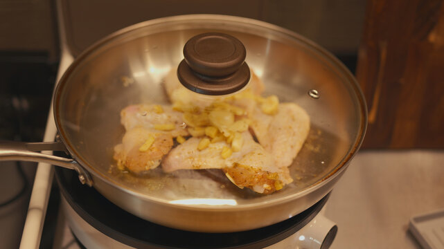 Sizzling chicken wings braising under a glass lid on a portable induction cooktop. Frying poultry meat with sliced ginger, minced garlic, and aromatic spices in a stainless steel pan in home kitchen.