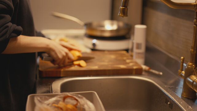 Selective focus close-up on brass faucet and kitchen sink with woman slicing peeled sweet potato with knife on wooden board in blurred background. Food prep process in cozy warm home kitchen