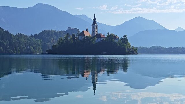 Bled Island and the Church of the Assumption on Lake Bled, Slovenia, surrounded by calm blue water and Alpine mountains, with reflections and sunlight visible on the lake.