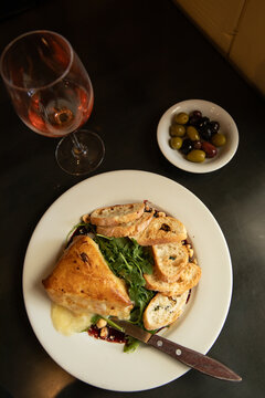 overhead shot of arugula salad with baked brie and crostini and side of olives and glass of wine