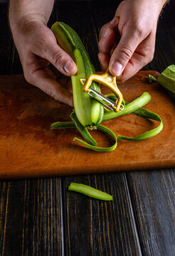 Zucchini peeling continues on cutting board as hands guide yellow peeler over fresh squash. Tight food prep detail shows green curls, steady grip, and concentrated home cooking moment.