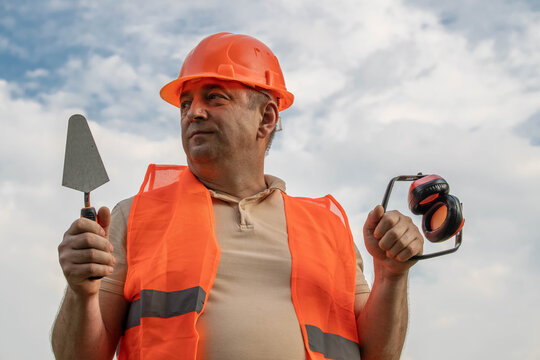 A construction worker stands outside holding a trowel in one hand and ear protection in the other. He wears a bright orange vest and helmet. The sky is mostly cloudy