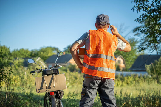 A delivery worker in a bright orange vest stands beside his bicycle, holding a box while talking on his phone. Lush greenery and homes dot the landscape under a clear blue sky