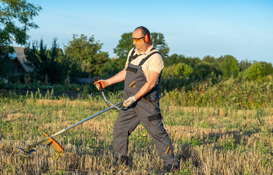 Worker using brush cutter on harvested field, standing with sturdy stance and protective gear. Man clears weeds and straw with careful control, showing practical outdoor labor and focused effort.