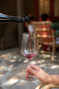 female hand holding a wine glass receiving a pour of red wine at an outdoor winery, resting hand on a rattan arm chair