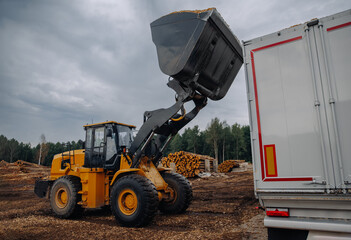 Fototapeta premium Yellow front-end loader lifting a bucket of wood chips above a white truck at a lumber yard with stacks of logs and cloudy sky in the background