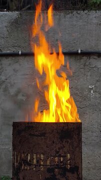Rusty metal barrel with an open fire burning inside, standing against a weathered wall outdoors, with bright orange flames and dark smoke rising from the barrel.