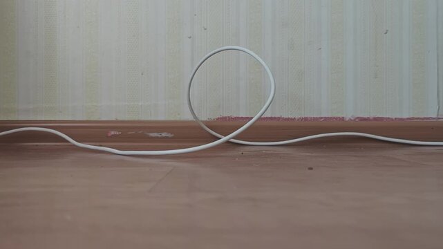 Looped white electric wire twisting and turning on a dusty wooden floor. Shabby room with worn wallpaper and a baseboard in the background
