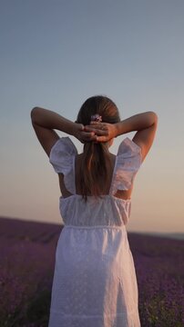 Lavender field serenity captures a young woman tying her hair in a white dress at dusk. Vertical medium shot.