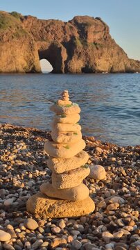 Stacked stone cairn stands on a pebble beach with a rocky arch in the background. Steady handheld vertical shot.