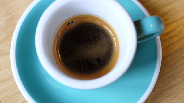 Blue espresso cup on wooden table stays centered as hand steadies saucer, showing rich coffee surface. Espresso drink remains ready for serving in calm cafe setting, evoking quiet morning pause.