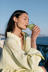 Fototapeta premium Profile of a woman sipping green juice with ocean view in an oversized yellow shirt for summer wellness lifestyle and travel banner