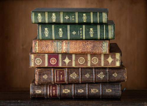 Stack of antique books with richly decorated leather bindings, gilt tooling, raised bands and ornate embossed spines, displayed against a warm wooden background. 