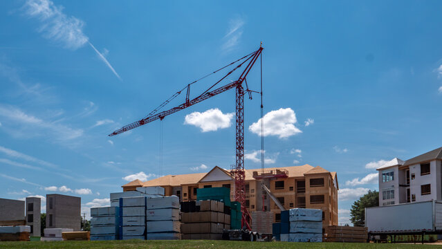 A construction site is shown with a large crane lifting materials. There are stacks of building supplies in the foreground. The sky is clear with a few clouds. Nearby buildings are under development.