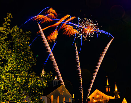 Fireworks explode in the night sky, creating bright colors and patterns above a town. Trees and buildings are visible below, providing a backdrop to the celebration. The atmosphere is festive.