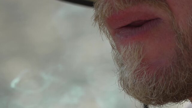Bearded man smoking handmade cigar with wooden splint holder outdoors near beach. Close-up macro tracking shot.