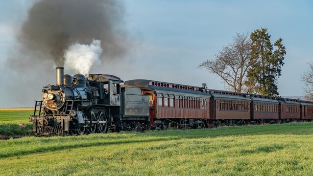 A steam train moves along tracks surrounded by green fields and trees. Smoke rises from the train as it approaches. The sky is clear and bright, capturing the moment of travel.