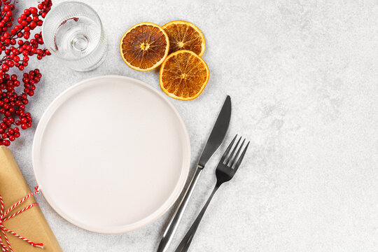 Christmas table setting with empty white plate, silver cutlery, red holly berries, dried orange slices, glass and wrapped gifts on light background. Template, mockup with festive holiday atmosphere.
