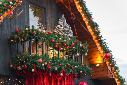 Festive Christmas wooden chalet balcony decorated with shining lights, illuminated evergreen garlands, ornaments, ribbons and a small tree, cozy winter holiday atmosphere with rustic architecture.