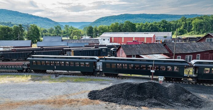 A vintage train moves through a rural town. The scene shows green hills in the background and several old buildings nearby. The train connects different parts of this quiet area.