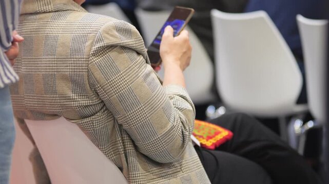 Woman sits in chair, holding phone. She wears checkered blazer. White chairs line background. Her phone screen glows blue. Red patterned bag rests beside her. Scene shows indoor event seating