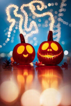 Two glowing Halloween jack-o'-lantern pumpkins with cute and scary carved faces, beautiful autumn decoration with teal background, fairy light bokeh, and reflections