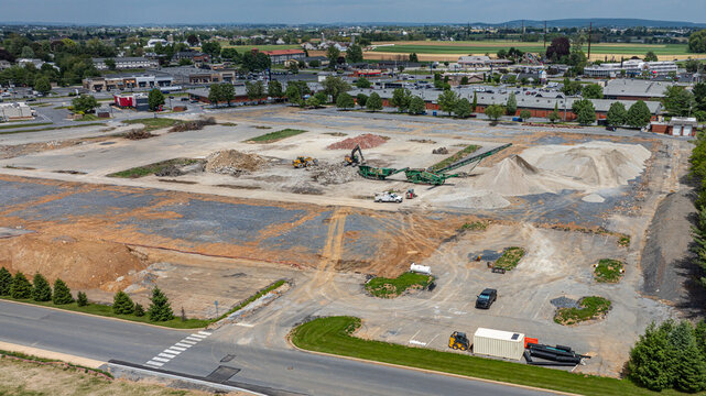 A construction site shows heavy machinery working to move dirt and materials. The area is rural with fields in the background. It is bright and the sky is clear.