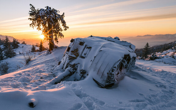 Overturned military tank on snowy mountain at sunset with snow and tree, landscape, war concept.