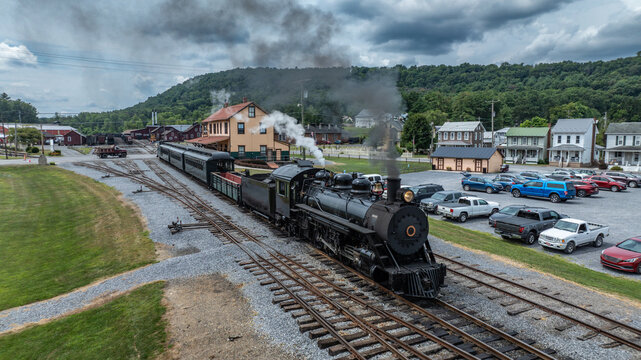 A large steam locomotive is at a train station with multiple train cars. Smoke billows from the engine. Several parked cars are visible. Green hills are in the background. It is daytime.