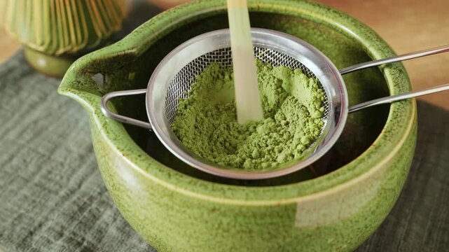 Pressing and sifting bright green matcha powder through metal mesh sieve into green ceramic bowl chawan with bamboo wooden spatula scoop chashaku. Traditional Japanese matcha tea preparation.