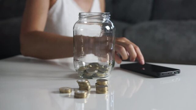 Close-up shot of a woman arranging stacks of euro coins on a table and dropping coins into a transparent glass savings jar.
