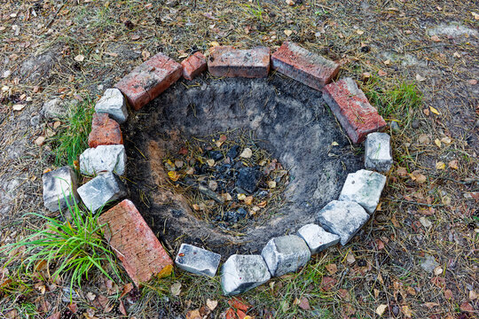 Rustic circular fire pit constructed from aged red bricks and stone blocks nestled in a forest clearing with dry pine needles and natural dirt ground for outdoor camping adventures