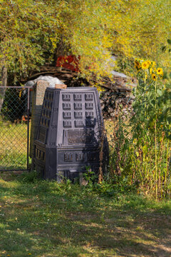 Black plastic garden composter in a sunny backyard corner next to a wire fence and tall blooming sunflowers for recycling.