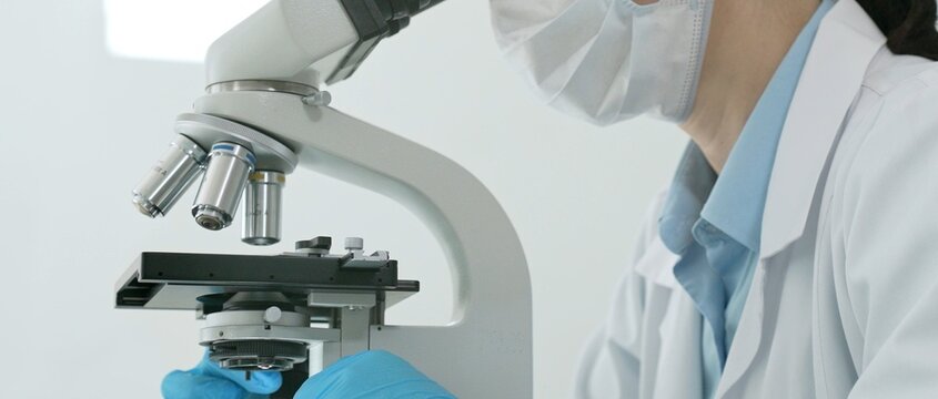 Scientist wearing face mask and protective glasses, looking through a microscope, working on medical research in a clean laboratory setting, analyzing biological samples