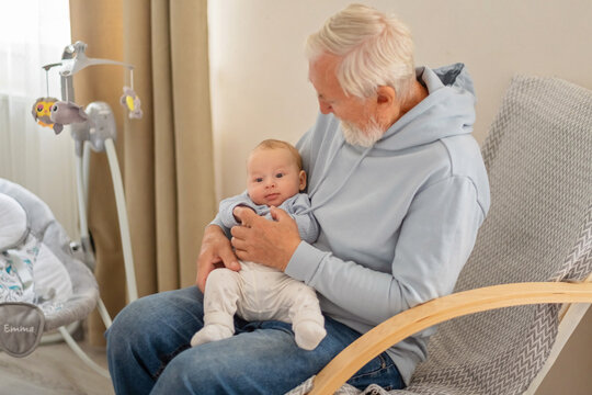 Elderly man sits with baby in nursery beside electric swing, generation gap turning into warm contact, mood for family campaign. Wide shot. Concept of generation passing care at home