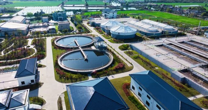 High angle view of a modern sewage treatment plant featuring large circular clarifier tanks and blue roof buildings under bright daylight.